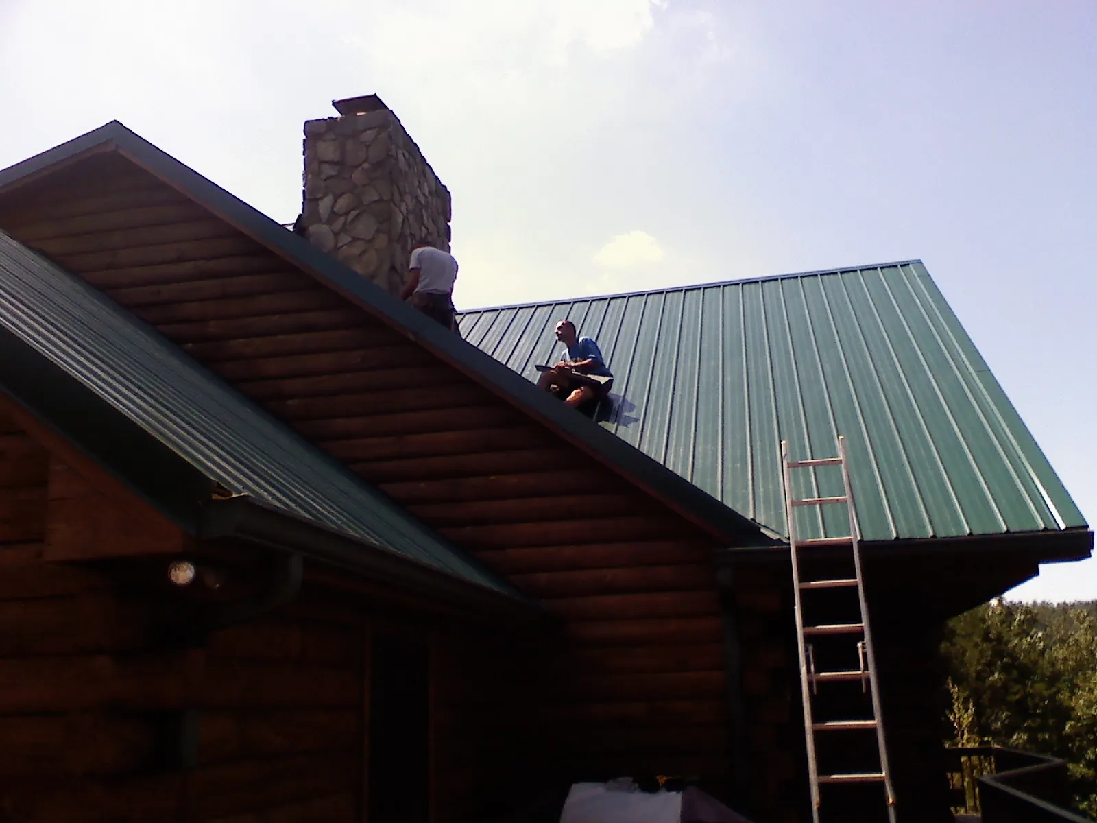 Roofers installing green metal panels beside a stone chimney on a log home in Brookfield